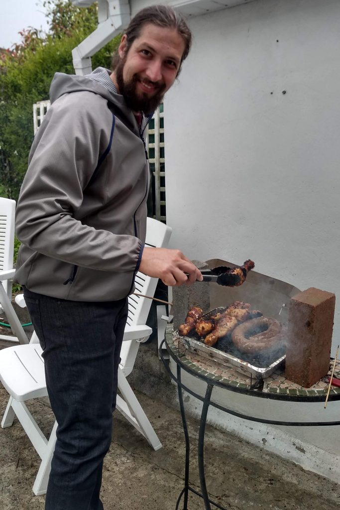 A man wearing a light raincoat standing outside in grey weather barbecuing pieces of chicken and a coil of sausage over an aluminium tray of coals. A brick and some sheets of metal are propped up to protect the barbecue from the weather.